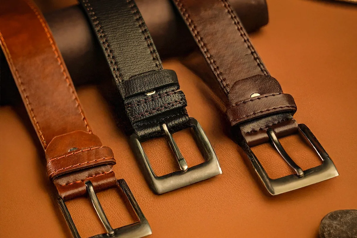 Three brown and black leather belts with metal buckles lying side by side on a tan surface